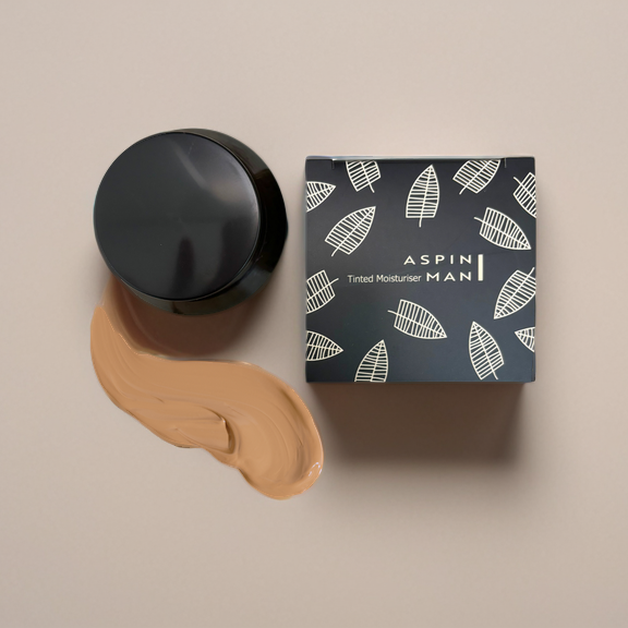 Top-down view of the Aspin Man Tinted Moisturizer jar, its sleek black and white packaging box, and a smooth swatch of the tinted moisturizer on a beige background, showcasing the product’s texture and branding.